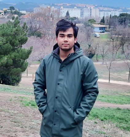 Picture of Sheikh with city buildings behind him and trees wearing a green coat with a hood, hands in the pockets.